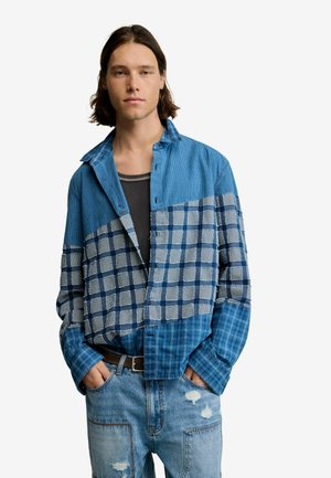 Młody mężczyzna z włosami sięgającymi ramion, ubrany w niebieską koszulę w kratę z naszywkami na czarnym tank topie i podniszczonych dżinsach, z rękami w kieszeniach.