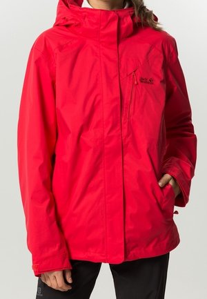 Femme portant une veste imperméable rouge vif avec capuche, poche poitrine zippée et poches latérales, debout devant un fond uni.