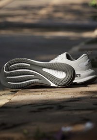 Baskets blanches allongées sur le côté à l'extérieur, montrant une semelle texturée noire et blanche avec des motifs incurvés sur une surface en pierre.