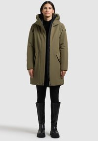 Oliwkowa parka z kapturem, z zamkiem błyskawicznym z przodu, dwoma bocznymi kieszeniami i regulowanymi mankietami, noszona na czarne prążkowane legginsy i wysokie buty do kolan.