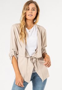 Beige, lichtgewicht wikkeljack met lange mouwen, open voorkant, een strik in de taille en opgerolde manchetten, gecombineerd met een witte t-shirt en blauwe spijkerbroek.