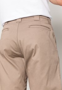 Pantalons beiges avec une texture lisse, dotés de deux poches arrière et de passants de ceinture. Le matériau semble robuste et durable.