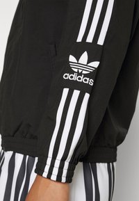 Svart jacka med vitt Adidas-logotyp och tre vertikala ränder på ärmarna. Materialet är slätt med elastiska mudd.