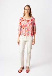 Blusa a fiori nei toni di rosa, rosso e blu, con maniche 3/4, abbinata a pantaloni bianchi a gamba stretta e sandali rossi, su uno sfondo semplice.