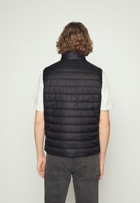 Czarna pikowana kamizelka z wysokim kołnierzem, ozdobiona poziomymi przeszyciami. Nosi się ją na białej koszulce, w połączeniu z szarymi spodniami.