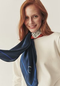 Sciarpa di seta blu marin con bordo blu e logo, indossata intorno al collo, abbinata a un maglione bianco. Texture liscia con caduta fluida.