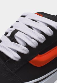 Gros plan sur une basket noire avec des lacets blancs et une large bande orange sur le côté, mettant en valeur le laçage et les matériaux texturés.