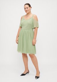 Lichtgroene chiffon jurk met cold shoulder design, uitlopende rok en geplooide textuur. G gedragen met zwarte puntige slingback schoenen.