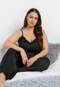 Kvinna med långt brunt hår som sitter på sängen, klädd i en svart satinkimono med spetskant, med en grå texturerad kudde och en stickad filt i närheten.