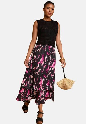 Fekete kötött crop top ujjatlan kialakítással, amelyet egy fekete, rózsaszín és fehér árnyalatokban pompázó, réteges, hosszú virágmintás szoknyával párosítanak. Szalmából készült legyezőt tart.