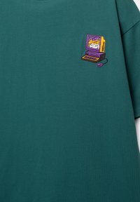 Zöld pamut póló, amelyen egy kis hímzett mintázat található egy vintage számítógépről színes díszítésekkel, bal mellrészen elhelyezve.