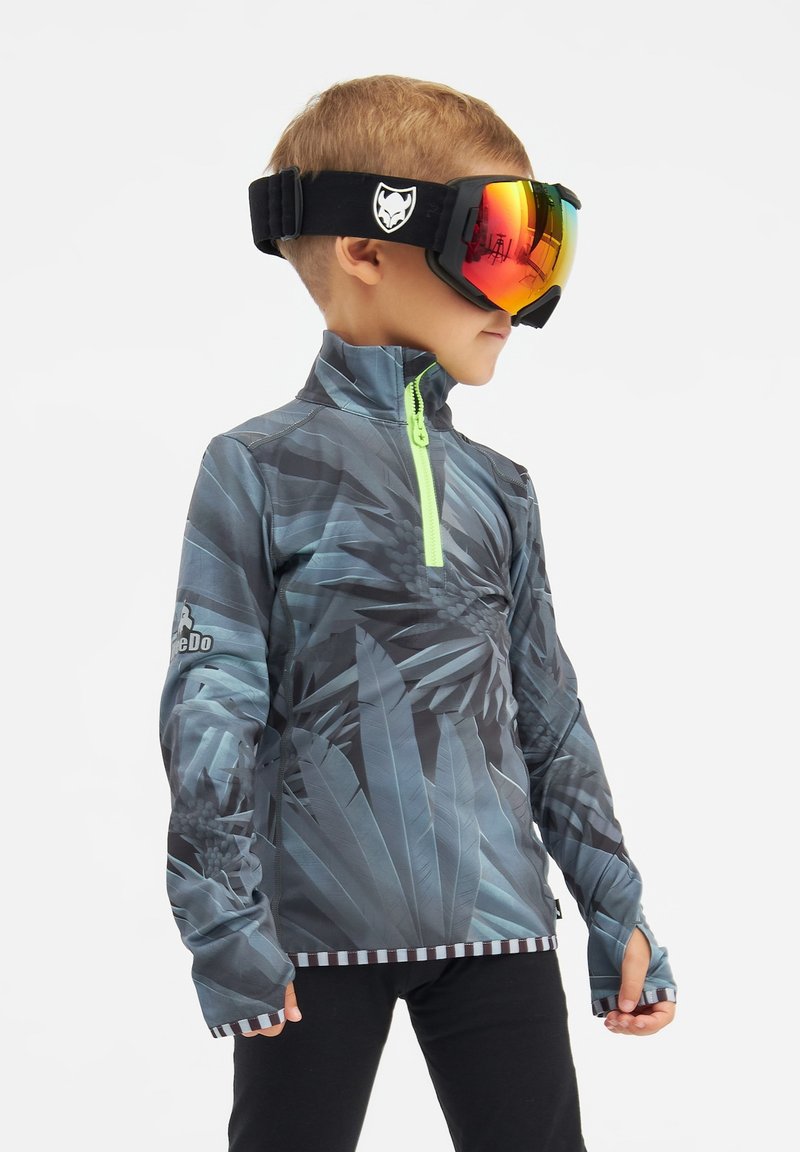Camiseta de manga larga con cremallera, que presenta un patrón de hojas tropicales oscuras, base gris-azulada y cremallera verde neón. Combinada con gafas de esquí espejadas.