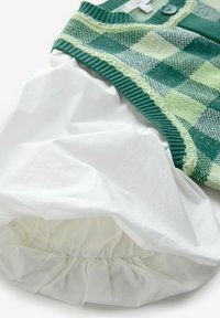 Zelená kostkovaná pletená vesta s žebrovaným límcem, spárovaná s bílým nařaseným spodním dílem. Textura je měkká a vrstvená.