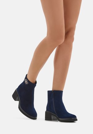 Stivaletti alla caviglia in suede blu navy con tacco spesso, chiusura laterale a cerniera e dettagli decorativi in argento sulla caviglia. Texture liscia, design aderente.
