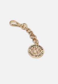 Klíčenka ve zlatém tónu s karabinou, zdobená kulatým přívěskem s raženým nápisem "Donna Karan" a "Circa 1989" černým písmem.