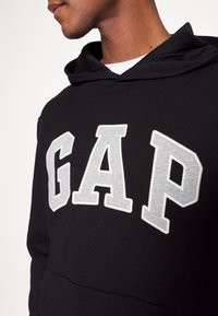 Sudadera negra con el logotipo "GAP" bordado en gris, con bolsillo tipo canguro y puños acanalados. Material de algodón suave con un corte casual.