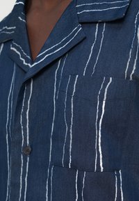 Μπλε πουκάμισο με λευκές κάθετες ρίγες, κλείνει με κουμπιά και έχει τσέπη στο στήθος. Κατασκευασμένο από υφασμάτινο ύφασμα με σχέδιο γιακά.