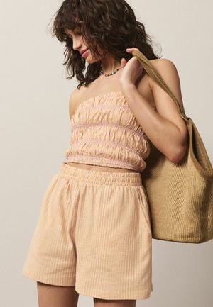 Femeie cu păr creț și închis la culoare, purtând un top piersică fără bretele cu detalii drapate și pantaloni scurți asortați, ținând o geantă mare țesută de culoare bej pe umăr.