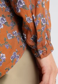 Langsleeve blouse in verbrande sinaasappel stof met paars bloemenpatroon, voorzien van een knoopsluiting bij de mouwen en een lichte, gestructureerde afwerking.
