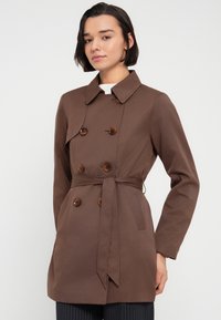 Καφέ διπλής σειράς παλτό με ζωνάκι στη μέση, διακοσμητικά κουμπιά και κλασικό γιακά. Κατασκευασμένο από απαλό ύφασμα με εφαρμοστή γραμμή.