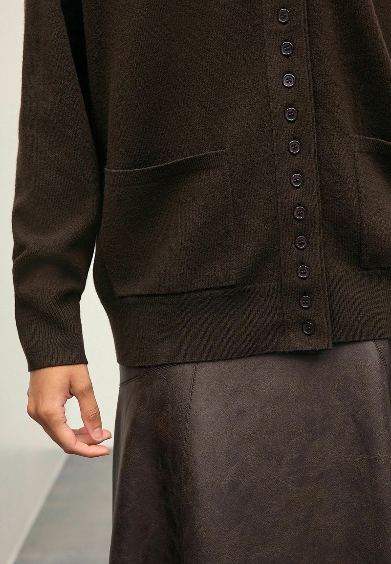 Gros plan d'une personne portant un cardigan noir boutonné avec des poches et une jupe en cuir marron foncé, main détendue le long du corps.