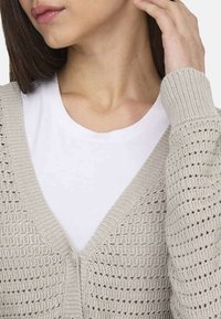 Cardigan en maille beige clair avec un décolleté en V et un motif texturé, superposé à une chemise blanche, mettant en valeur le tissage ouvert de la maille.
