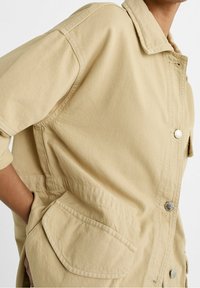 Béžová bavlněná bunda s límcem, stříbrnými patenty, bočními kapsami a uvolněným střihem. Obsahuje texturovaný povrch a viditelné stehy.