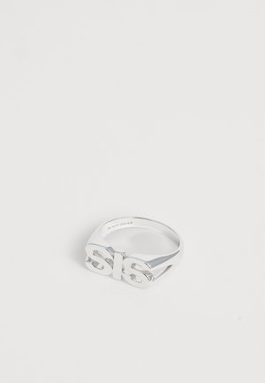Sølvmetal ring med fede, hævede bogstaver "sls" på toppen, sat mod en ensfarvet hvid baggrund.