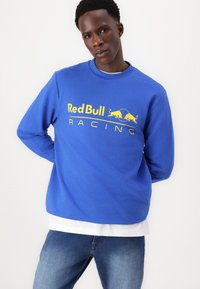 Modrá mikina vyrobená z bavlněné směsi, na přední straně s velkým žlutým logem "Red Bull Racing" a žebrovaným kulatým výstřihem.