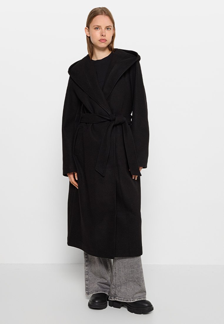 Svart ullsjal med bred krage, frontknut och långa ärmar, bärs över grå vida byxor och klumpiga svarta skor.