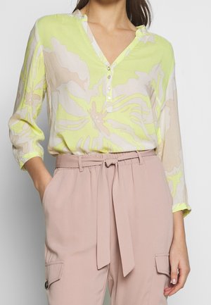 Woman wearing a light yellow floral blouse with three buttons and beige high-waisted pants with a tied belt and pockets.