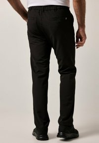 Zwarte, stretch stof broek met een elastische tailleband, achterzakken met knoopsluitingen en een taps toelopend ontwerp. G gedragen met zwarte sneakers.
