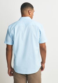 Mann trägt ein hellblaues Kurzarmhemd und braune Hose, steht mit dem Rücken zu einer schlichten weißen Wand.