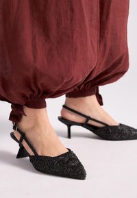 Tacones de gatito negros con tiras traseras y patrones florales texturizados, usados con pantalones holgados y recogidos de color óxido, atados en los tobillos.