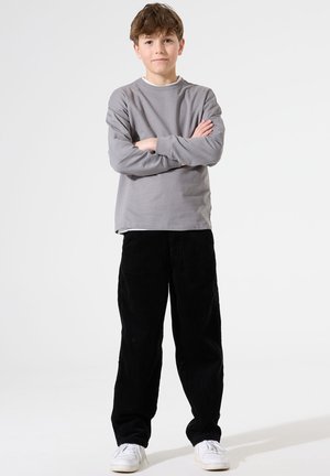 Graues Langarmshirt mit Rundhalsausschnitt, kombiniert mit schwarzen Cordhosen und weißen Turnschuhen. Kind steht mit verschränkten Armen.