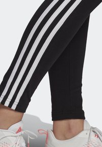 Leggings neri con tre strisce bianche sui lati, abbinati a sneakers bianche con dettagli rosa. Inquadratura ravvicinata.