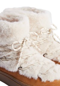 Bottines fourrées à la cheville avec une tige crème douce et des accents en suède marron, dotées de lacets crème avec un motif marron subtil et de crochets en métal.