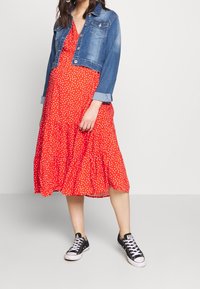 Chaqueta de mezclilla con botones en la parte delantera, mangas largas y puños enrollados, combinada con un vestido rojo con lunares que presenta un dobladillo con volantes, usado con zapatillas deportivas negras.