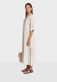 Robe maxi en lin beige clair avec manches courtes, décolleté plongeant en V, bordure noire contrastante, fentes latérales, portée avec des sandales noires.