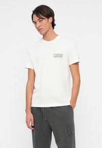 Fiatal férfi fehér grafikus pólóban és sötétszürke, hímzett feliratos melegítőnadrágban áll, egy kezét a zsebébe téve fehér háttér előtt.