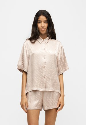 Femme aux longs cheveux foncés portant une chemise en satin rose clair à manches courtes boutonnée et un short assorti, debout devant un fond uni.