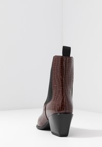 Botín de cuero sintético color marrón con un panel lateral elástico negro y un tacón afilado. Presenta una pestaña en la parte posterior para facilitar su uso.
