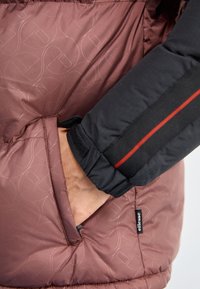 Chaqueta de plumas con un frente burdeos que presenta un patrón en relieve, mangas negras con detalles rojos y un bolsillo lateral. Se aprecian materiales texturizados.