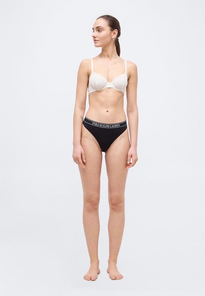 Γυναίκα που στέκεται ξυπόλυτη, φορώντας λευκό δαντελένιο σουτιέν και μαύρο εσώρουχο με το λογότυπο Polo Ralph Lauren, κοιτάζει προς τα αριστερά της με απλό φόντο.