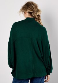 Person mit langen geflochtenen Haaren, die einen lockeren dunkelgrünen Pullover und blaue Jeans trägt, vor einer schlichten hellen Wand steht.