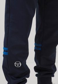 Temno modri trenirko hlače iz gladke tkanine, z tremi horizontalnimi svetlo modrimi črtami in belim logotipom na spodnjem delu noge.