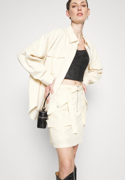 Veste surdimensionnée en velours côtélé beige, crop top en cuir noir, jupe crème avec poches et bottes noires hautes jusqu'aux genoux. Petit sac à main noir inclus.