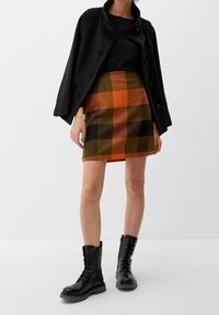 Zwarte cropped jas over een zwart topje, gecombineerd met een geruite minirok in oranje, groen en zwart. Zwarte veterschoenen.