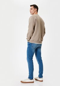 Felpa beige con maniche lunghe, scollo rotondo e polsini a coste. Indossata con jeans blu e sneakers bianche, vista da dietro.