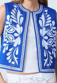Blå vest med indviklet hvid blomsterbroderi, rundet halsudskæring, åben front og dekorativ kant langs kanterne.